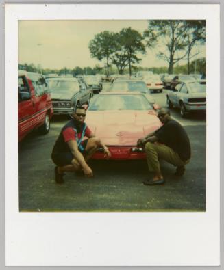[Two men kneeling in front of red car in parking lot]

