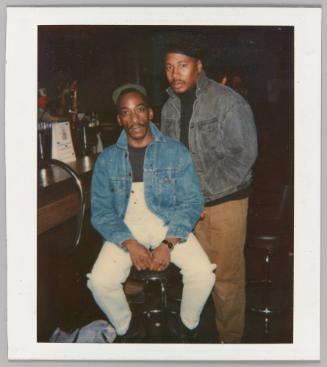 [Two men at barside; man on left wearing denim overalls]
