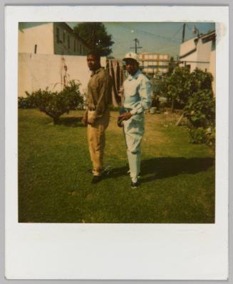 [Two men posing outside on grass; man on right wearing Boston Red Sox baseball cap]
