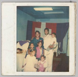 [Man, woman, three little girls, 1 little boy and a toddler posing in front of flag]
