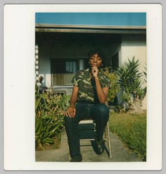 [Woman on seated on chair outside a home with tropical plants wearing a camouflage shirt]
