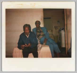 [Two elderly men sitting on chair covered by a bedsheet; man in background smiling and holding drink]
