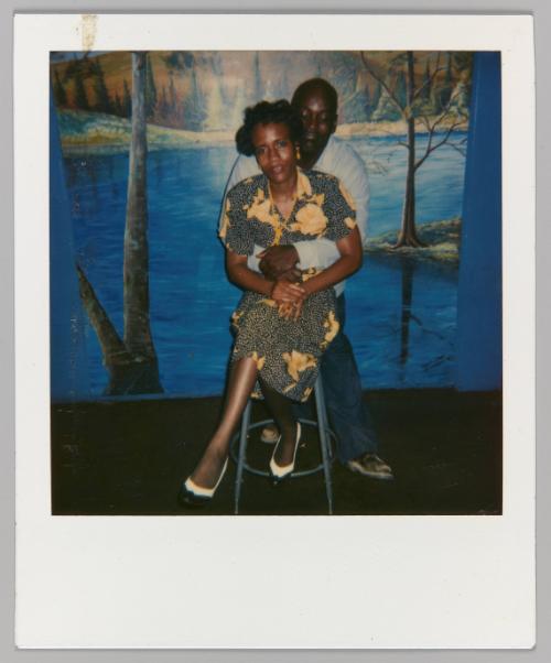 [Man embracing seated woman around the waist from behind; posing in front of landscape mural]

