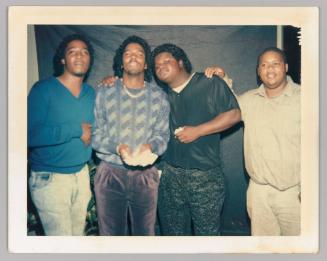 [4 men pose together holding each others shoulders; man second on the left holding a stack of money]
