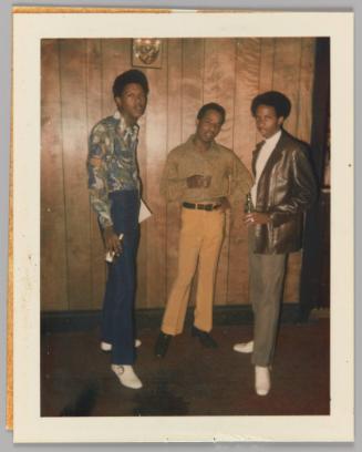 [Three men standing in front of wood panel wall holding beers]
