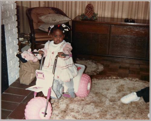 [Young girl with hair bows and dress sitting on white and pink tricycle, she is standing on a shag rug and in front of a white brick fireplace]