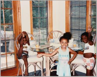 [Three girls posing by glass dining table surrounded by windows with wood frames, girl on left is sitting and holding her foot up in the air]