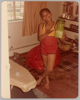 [Boy in red and yellow athletic wear posing with a "Hulk" statue, behind is a red beanbag chair and a shelf with trophies]