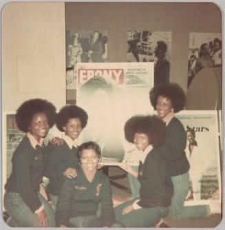 [Five women, four with afros and matching collared shirts and sweaters, posing indoors and holding up a poster that reads "Ebony"]