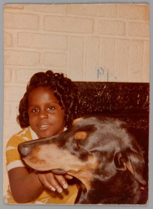 [Girl with curled hair and a dog in front of white brick fireplace, she has her hand beneath the dog's snout]