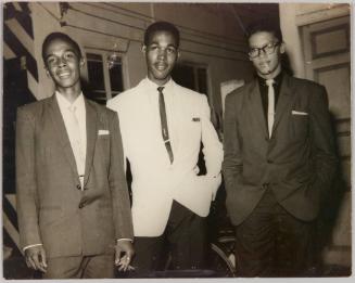 [Three men standing inside in suits, man in centre wearing white jacket and man on right has a cigarette in his mouth]