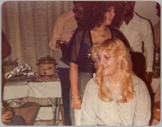 [Woman with blonde layered hair sitting down, there are other people and a table with food]