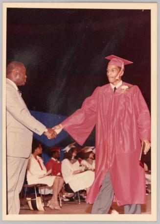 [Young man in red graduation gown and cap on stage and shaking hands with man in suit, there are people sitting down behind]