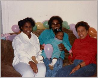 [Three women sitting on couch, woman in centre holding a young girl with pink balloon, colourful balloons behind couch]