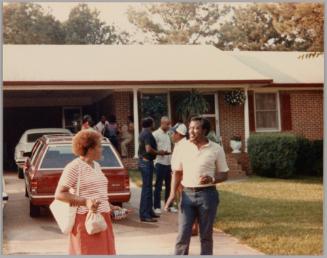 #19 [Woman in red and white striped shirt and man holding glass in front of people standing on front lawn and driveway of house]