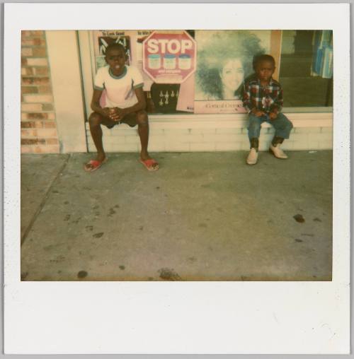 [Two boys sitting outside on a ledge in front of a store window]