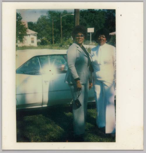 [Two women standing in front of car, woman on left in pantsuit and woman on right in white outfit]