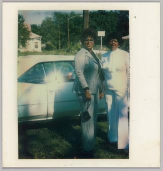[Two women standing in front of car, woman on left in pantsuit and woman on right in white outfit]