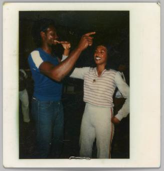 [Smiling woman standing with one hand on hip and other hand raised, with man pointing to the right ]