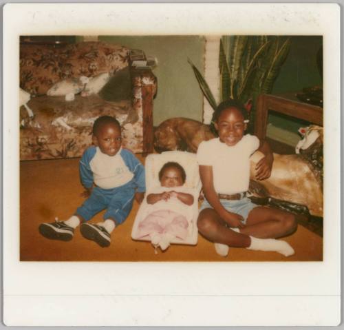 [Boy and girl sitting on floor with baby in white carrier between them]