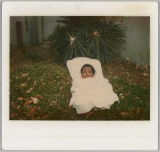 [Baby in white carrier and blanket on lawn with fallen leaves, fence behind]
