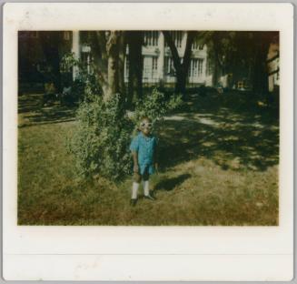 [Child in large sunglasses standing on lawn in front of bush and trees]