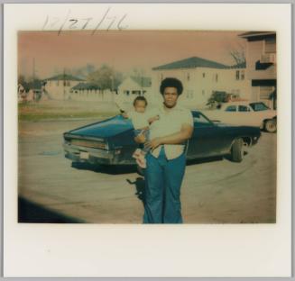 12/27/76 [Man holding baby in one arm, he is standing in a parking lot in front of a black car]