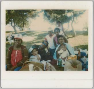 [Group of people posing around picnic table with food, woman to far right is holding a baby]