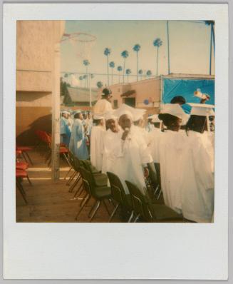 [Children standing in white gowns and caps at outdoor event, basketball net to the left]