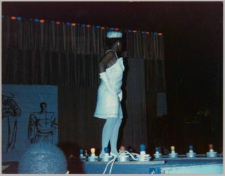 [Woman on stage in white dress, tights, and hat with elbow-length gloves]