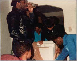 [Profile of man dressed in black standing and drinking from a glass with people bending down and opening package]