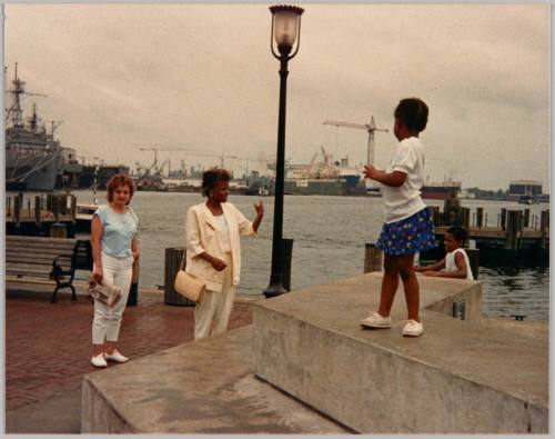 #1 [Two women standing on boardwalk by water with two children, there is a ship to the left]