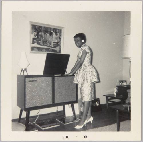 [Woman in metallic silver patterned outfit standing by record player]
