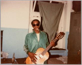 [Man with cigarette hanging from his mouth wearing cowboy hat and sunglasses, holding guitar]
