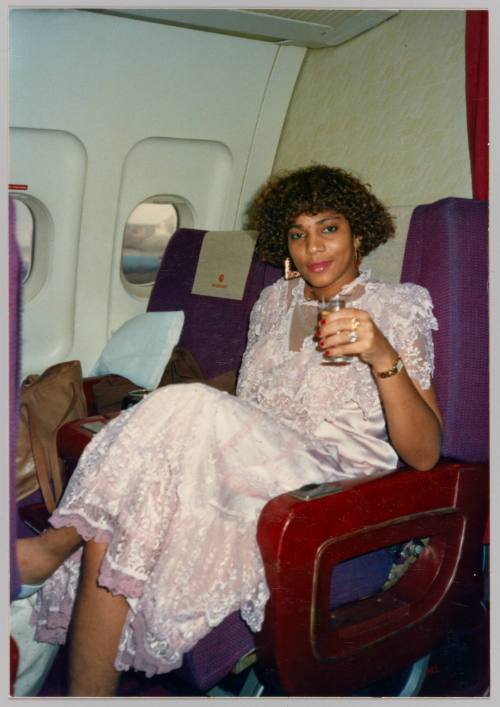 [Woman in pink lace dress with drink in hand seated on an airplace]

