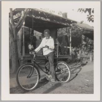 [Child smiling and mounting bicycle in front of porch]
