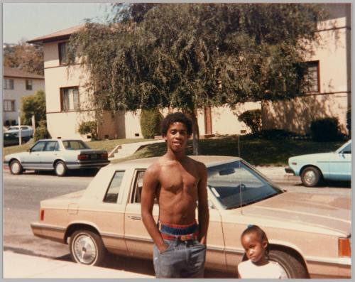[Adolescent man with bare chest standing with hands in pockets in front of street with little girl at his side]
