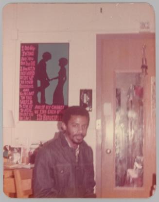 [Man sitting in room with mirror on door and textual poster]
