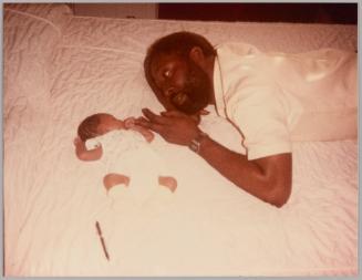 [Man laying on bed with baby holding his middle finger in fist]
