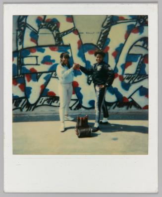 [Two young women posing in front of a graffiti wall giving each other a fist-bump]
