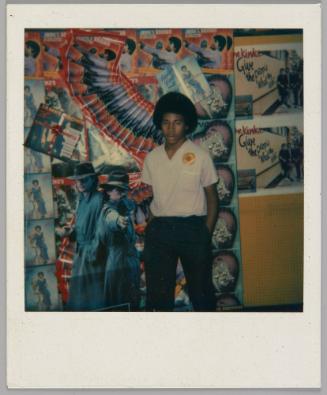 [Teenage boy in white v-neck t-shirt and black pants with his hands in pockets standing by wall covered in music posters, including James Brown and The Kinks]