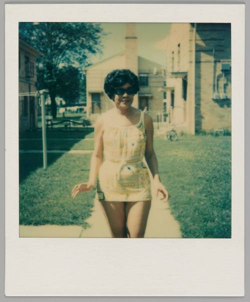 7-16-78 Thelma 53 Taken by Judy [Woman with short hair wearing sunglasses and a short outfit that has a tie at the waist, she is standing on a walkway between grass lawns]