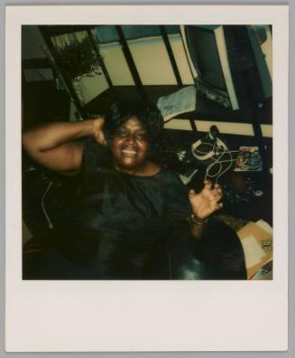 [Woman sitting in front of television set and smiling up at camera, she has one hand behind her head and the other hand formed as a claw]