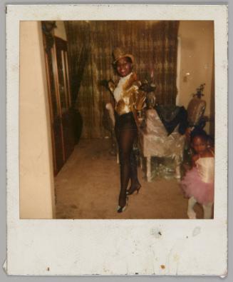[Woman posing inside in show girl outfit with a gold hat, gold jacket, and black stockings, girl in pink ballerina outfit to right]