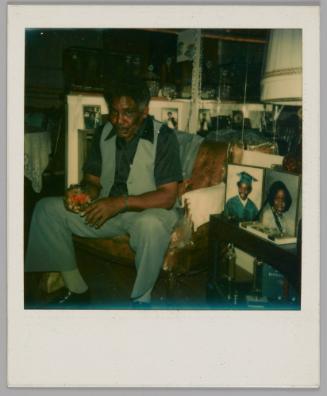 [Man wearing an open vest and blue short-sleeved shirt and sitting in armchair with a drink in his hand, he is surrounded by family photographs]