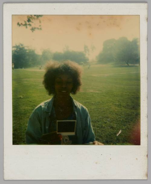 [Woman holding and looking down at polaroid in a park with a large empty field, she is wearing an unbuttoned blue shirt]