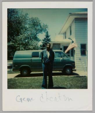 Gene Cheaton [Man in jacket and flared pants standing with his hands behind his back, he is on lawn in front of a driveway with parked blue van and an American flag]
