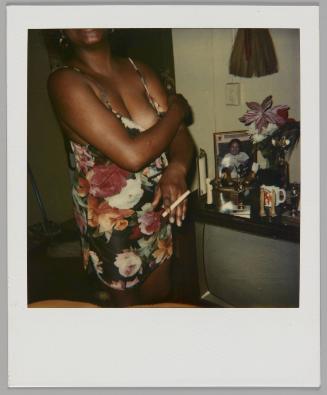 [Woman wearing a sleeveless floral dress with unlit cigarette in one hand and other hand on opposite shoulder, standing beside television set with framed photo, vase, and knick knacks]