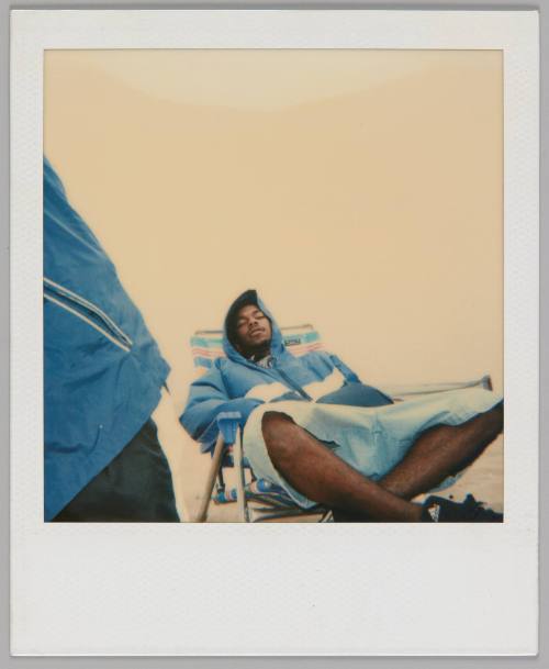 [Man in blue jacket and shorts reclining with his legs crossed on beach chair, someone's jacket visible on left]