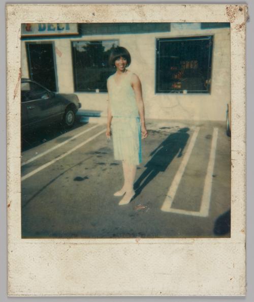 [Woman in light blue top and skirt and white stockings and shoes standing in the parking lot of a building with a "Deli" sign to the left]
